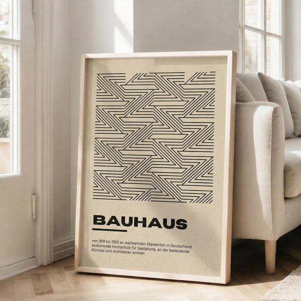 May include: Beige framed poster with black geometric line pattern and "BAUHAUS" text, leaning against white wall next to beige upholstered chair in bright room with large windows and light wooden floors