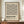 May include: Framed geometric poster with black linear pattern on cream background, featuring intersecting diagonal lines forming repeating triangular shapes. Bold "BAUHAUS" text below with German description. Displayed on wooden sideboard beside white ceramic vase and stacked books in neutral-toned room with white paneled walls.