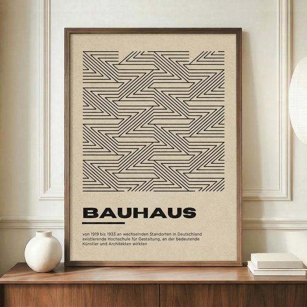 May include: Framed geometric poster with black linear pattern on cream background, featuring intersecting diagonal lines forming repeating triangular shapes. Bold "BAUHAUS" text below with German description. Displayed on wooden sideboard beside white ceramic vase and stacked books in neutral-toned room with white paneled walls.