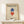 May include: Framed Bauhaus poster with geometric diamond shapes in blue and red on cream background, displayed on white console table next to wrapped gift with greenery. Poster features "Geometry and Simplicity" text and exhibition details from 1919. Light wooden frame complements minimalist room setting with white walls and natural lighting.