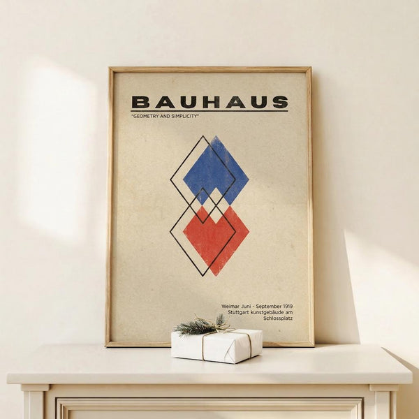 May include: Framed Bauhaus poster with geometric diamond shapes in blue and red on cream background, displayed on white console table next to wrapped gift with greenery. Poster features "Geometry and Simplicity" text and exhibition details from 1919. Light wooden frame complements minimalist room setting with white walls and natural lighting.