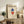 May include: Framed Bauhaus poster with geometric diamond shapes in blue and red on cream background, displayed on rustic wooden sideboard alongside fern plant, books, ceramic vase, and leather armchair in neutral-toned living room setting