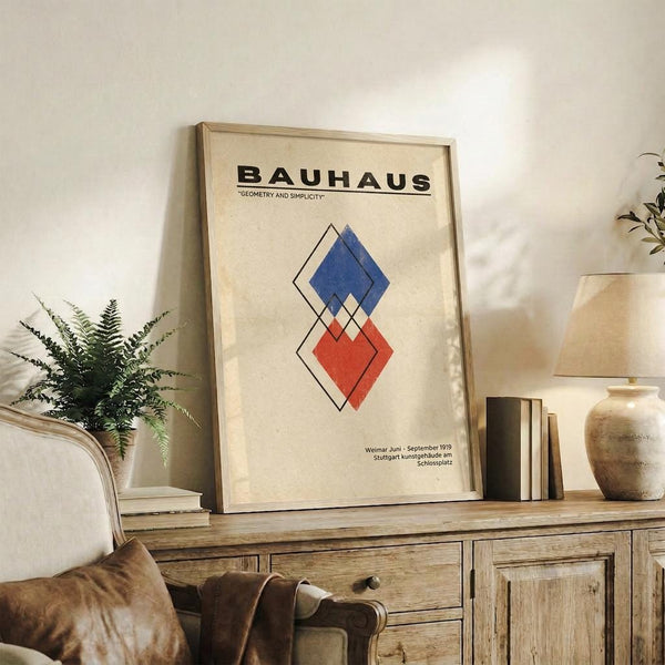May include: Framed Bauhaus poster with geometric diamond shapes in blue and red on cream background, displayed on rustic wooden sideboard alongside fern plant, books, ceramic vase, and leather armchair in neutral-toned living room setting