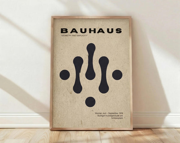 May include: A framed Bauhaus-inspired poster with geometric black shapes on cream textured paper, displayed against white walls on light wood flooring. The design features circles and elongated teardrop forms arranged symmetrically, with bold black text at top and smaller text at bottom. Natural wood frame with clean lines complements the minimalist aesthetic.