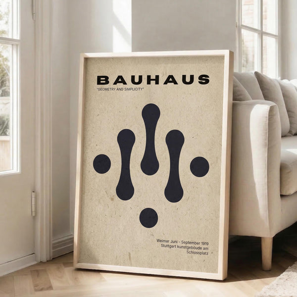 May include: Framed Bauhaus-inspired poster with cream textured background and black geometric shapes arranged symmetrically, featuring bold "BAUHAUS" text and "GEOMETRY AND SIMPLICITY" subtitle. Minimalist dark navy circles and elongated oval forms create balanced composition. German text at bottom references Weimar and Stuttgart locations with 1919 date. Light wood frame leans against white wall next to beige upholstered sofa in bright, airy room with large windows.