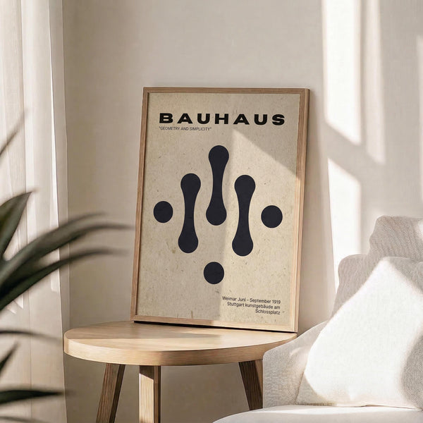 May include: Framed Bauhaus poster with black geometric shapes on cream background, displayed on light wood round side table next to beige upholstered chair. Natural sunlight streams across white walls, with green plant leaves visible in corner. The minimalist artwork features abstract droplet and circle forms in simple composition within thin wooden frame.