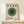 May include: Framed Bauhaus-style poster with concentric green circles in varying shades from light sage to deep forest green, four small circles across the top in matching tones, cream textured background, text reading "bauhaus" and "JUNE 17, 1921" at bottom, displayed in light wood frame on white wooden surface alongside small wrapped gift with natural twine and sprig decoration against white wall.