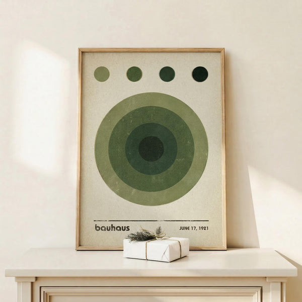 May include: Framed Bauhaus-style poster with concentric green circles in varying shades from light sage to deep forest green, four small circles across the top in matching tones, cream textured background, text reading "bauhaus" and "JUNE 17, 1921" at bottom, displayed in light wood frame on white wooden surface alongside small wrapped gift with natural twine and sprig decoration against white wall.