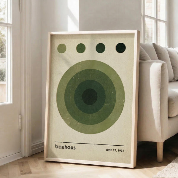 May include: Large framed Bauhaus-style poster featuring concentric green circles in varying shades from light sage to deep forest green, with four small circles above in graduated tones. The vintage-inspired print has "bauhaus" and "JUNE 17, 1921" text at bottom on cream background. Displayed in light wood frame leaning against white wall next to beige upholstered chair in bright, minimalist living room with large windows.
