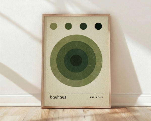 May include: Framed Bauhaus-inspired poster with concentric green circles in varying shades from light to dark, four small dots across the top in matching tones, cream background, and black text reading "bauhaus" and "JUNE 17, 1921" at bottom. Natural wood frame sits on light wooden floor against white wall with soft shadows.