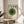May include: Framed Bauhaus-style poster with concentric green circles and four dots above, displayed on light wood round side table next to beige upholstered chair. Natural sunlight streams through white curtains creating geometric shadows on cream wall. Plant leaves visible in corner, creating minimalist modern interior styling with neutral tones.