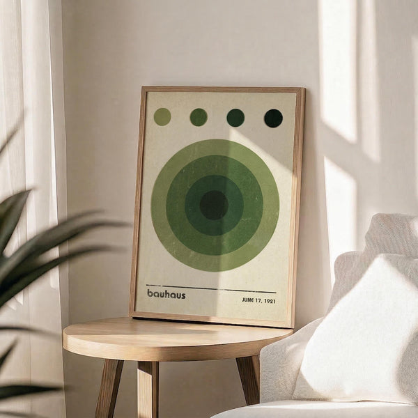 May include: Framed Bauhaus-style poster with concentric green circles and four dots above, displayed on light wood round side table next to beige upholstered chair. Natural sunlight streams through white curtains creating geometric shadows on cream wall. Plant leaves visible in corner, creating minimalist modern interior styling with neutral tones.
