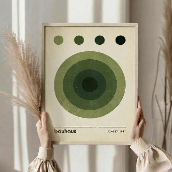 May include: Person in beige ruffled sleeves holding framed Bauhaus-style poster with concentric green circles and four dots above, natural wood frame, cream textured background, dried pampas grass visible on left side, soft natural lighting near white curtained window