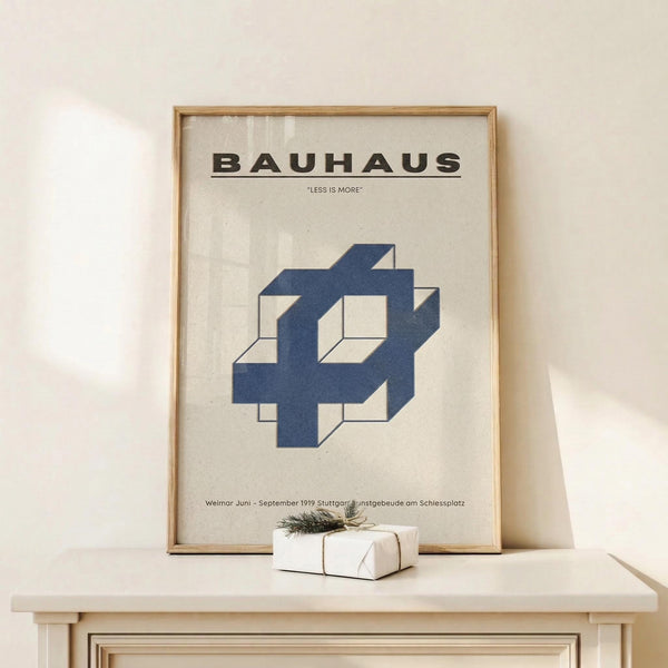 May include: Framed Bauhaus poster with blue geometric interlocking shapes and "LESS IS MORE" text displayed on light wood shelf against cream wall. Small wrapped gift box with twine and greenery sits beside the natural wood frame. Clean minimalist styling with soft natural lighting.