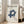 May include: Framed Bauhaus poster with cream background and navy blue geometric interlocking design, displayed on light wood floor against white wall corner. Beige upholstered sofa with cushions visible on right side. Natural sunlight streaming through tall white-framed windows creates soft shadows on wall. Poster features "BAUHAUS" and "LESS IS MORE" text with exhibition details below the geometric artwork.
