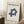 May include: Framed Bauhaus poster with cream background and navy blue geometric cube design, displayed leaning against white wall corner next to small wooden side table with white coffee cup and saucer, positioned on light wood floor with textured area rug and beige throw blanket visible in minimalist room setting.