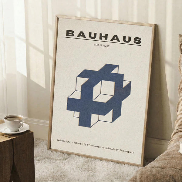 May include: Framed Bauhaus poster with cream background and navy blue geometric cube design, displayed leaning against white wall corner next to small wooden side table with white coffee cup and saucer, positioned on light wood floor with textured area rug and beige throw blanket visible in minimalist room setting.