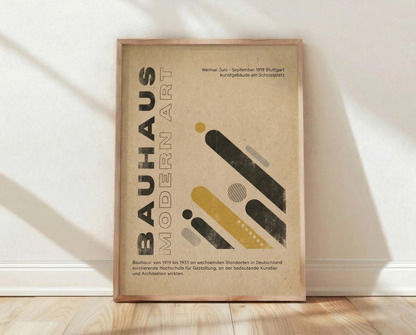 May include: Framed Bauhaus-inspired poster with geometric design featuring diagonal yellow and black bars, circles, and modern typography on beige background. Wood frame sits against white wall on light wooden floor with soft natural lighting creating shadows.