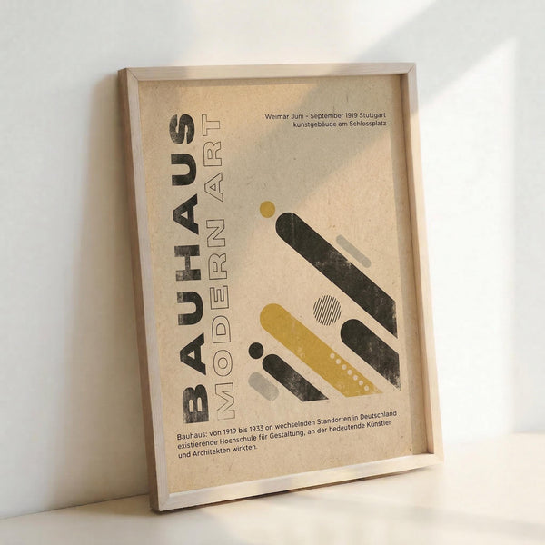 May include: Framed Bauhaus-inspired poster with geometric design in mustard yellow, dark green, and gray shapes on cream textured paper background. Modern art print displayed in light wood frame leaning against white wall, showing vintage typography and German text referencing the design school's history from 1919-1933.