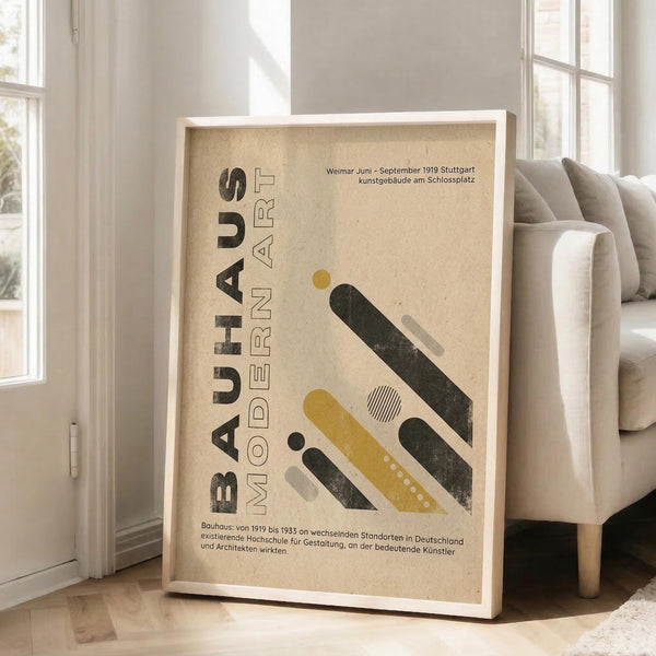 May include: Framed poster with beige textured background featuring geometric shapes in mustard yellow, dark green, and gray. German text reads "BAUHAUS MODERN ART" with historical information about Weimar and Stuttgart. White frame leans against white wall next to light gray upholstered chair in bright room with large windows.