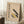May include: Framed Bauhaus poster with beige background featuring geometric shapes in yellow, black, and gray, German text about modern art movement. Poster leans against white wall next to white coffee cup on wooden block, cozy blanket visible, natural lighting creates soft shadows.