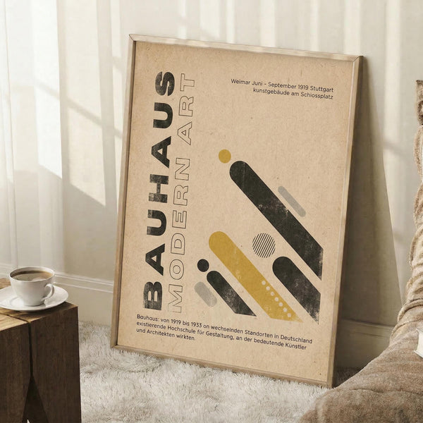 May include: Framed Bauhaus poster with beige background featuring geometric shapes in yellow, black, and gray, German text about modern art movement. Poster leans against white wall next to white coffee cup on wooden block, cozy blanket visible, natural lighting creates soft shadows.