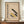 May include: Framed Bauhaus-inspired poster with beige background featuring geometric shapes in mustard yellow, dark green, and gray. German text reads "BAUHAUS MODERN ART" with exhibition details. Print displayed on wooden sideboard next to white ceramic vase and stacked books in modern interior with white paneled walls.