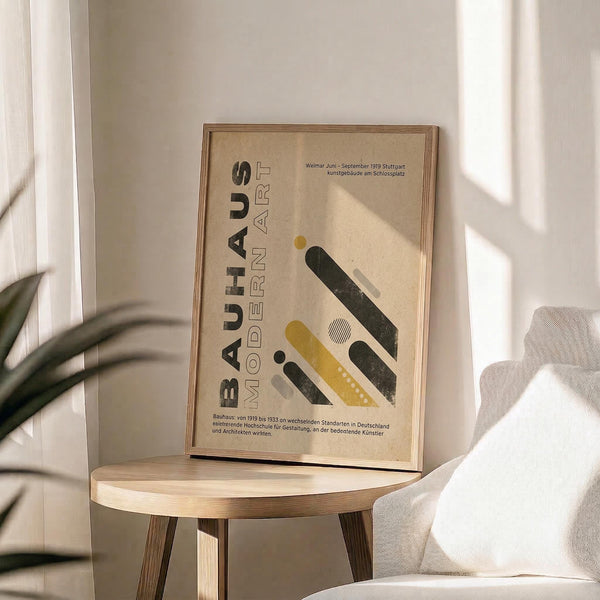 May include: Framed beige poster with "BAUHAUS MODERN ART" text and geometric shapes in black, yellow, and white displayed on light wood round side table next to cream upholstered chair. Sunlight streams through white curtains creating soft shadows. Green plant leaves visible in corner of minimalist room setting.