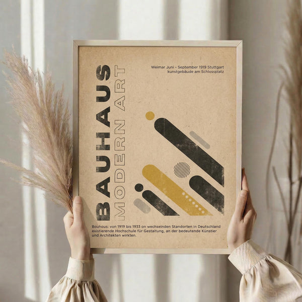 May include: Person holding framed Bauhaus-inspired poster with geometric shapes in mustard yellow, dark green, and cream colors on textured beige background. German text describes the art movement from 1919-1933. Natural light setting with dried pampas grass visible, wearing cream ribbed sweater sleeves.