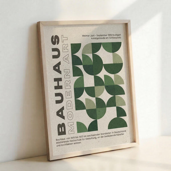 May include: Framed Bauhaus-style poster with geometric green circular and semi-circular shapes arranged in a grid pattern on cream background. Bold black text reads "BAUHAUS MODERN ART" vertically along left side. German text appears at top and bottom. Natural wood frame, displayed leaning against white wall with soft lighting creating subtle shadows.