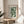May include: Framed Bauhaus poster with green geometric semicircle pattern and German text, displayed on light wood round table next to beige upholstered chair. Soft natural lighting creates shadows across white wall. Plant leaves visible in corner. Modern minimalist interior styling with neutral tones.