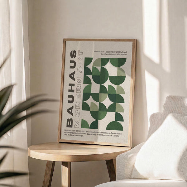 May include: Framed Bauhaus poster with green geometric semicircle pattern and German text, displayed on light wood round table next to beige upholstered chair. Soft natural lighting creates shadows across white wall. Plant leaves visible in corner. Modern minimalist interior styling with neutral tones.