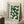 May include: Framed vintage-style poster with green geometric circles and semicircles arranged in a grid pattern, featuring "BAUHAUS MODERN ART" text and German exhibition details. Light wood frame leaning against white wall corner beside cream armchair, small wooden side table with white coffee cup, and textured cream rug.