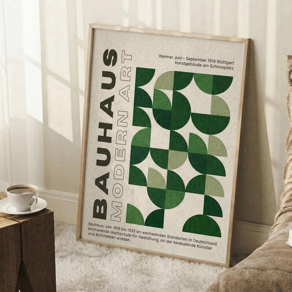 May include: Framed vintage-style poster with green geometric circles and semicircles arranged in a grid pattern, featuring "BAUHAUS MODERN ART" text and German exhibition details. Light wood frame leaning against white wall corner beside cream armchair, small wooden side table with white coffee cup, and textured cream rug.