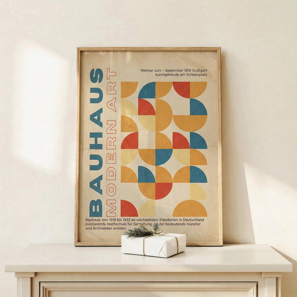 May include: Framed Bauhaus poster with geometric design featuring colorful circles and semicircles in orange, blue, red, and yellow arranged in a grid pattern. German text "BAUHAUS MODERN ART" appears vertically on left side. Wooden frame sits on light wood console table against white wall, with small wrapped gift beside it.