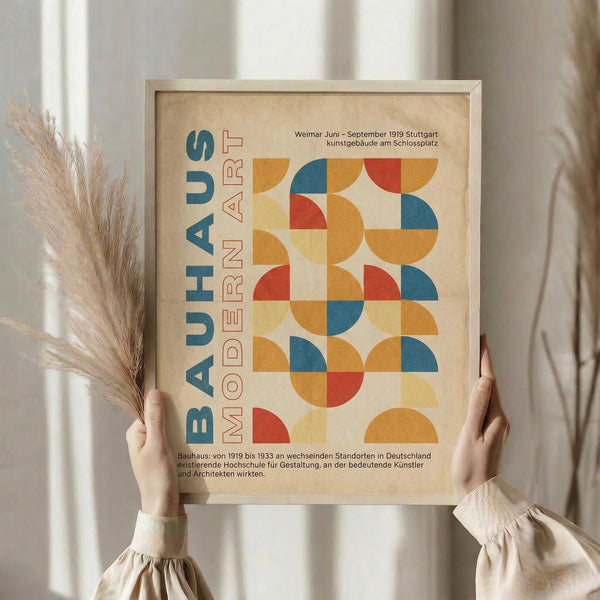 May include: Person holding framed Bauhaus-style poster with geometric pattern of orange, blue, and red semicircles and quarter-circles on cream background. German text reads "BAUHAUS MODERN ART" vertically. Light wood frame, soft natural lighting, dried wheat stems visible on left, neutral beige clothing and curtains in background.