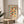 May include: Framed vintage-style Bauhaus poster with geometric semicircles in orange, blue, red and yellow, displayed on round wooden side table next to beige upholstered chair. Natural sunlight streams through white curtains creating soft shadows. Green plant leaves visible in corner. Poster features "BAUHAUS MODERN ART" text and German exhibition details on cream background in thin wooden frame.