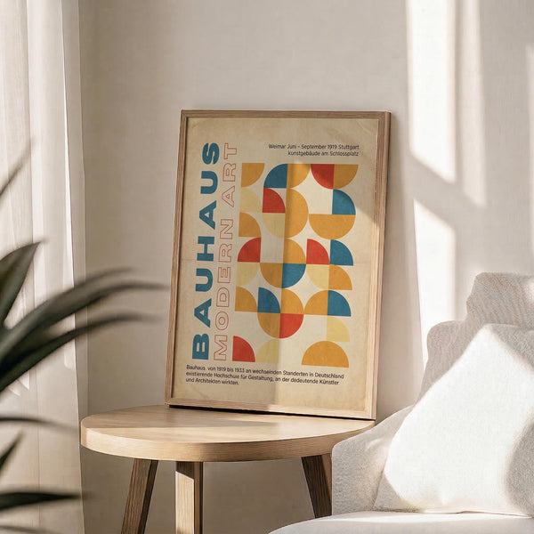 May include: Framed vintage-style Bauhaus poster with geometric semicircles in orange, blue, red and yellow, displayed on round wooden side table next to beige upholstered chair. Natural sunlight streams through white curtains creating soft shadows. Green plant leaves visible in corner. Poster features "BAUHAUS MODERN ART" text and German exhibition details on cream background in thin wooden frame.