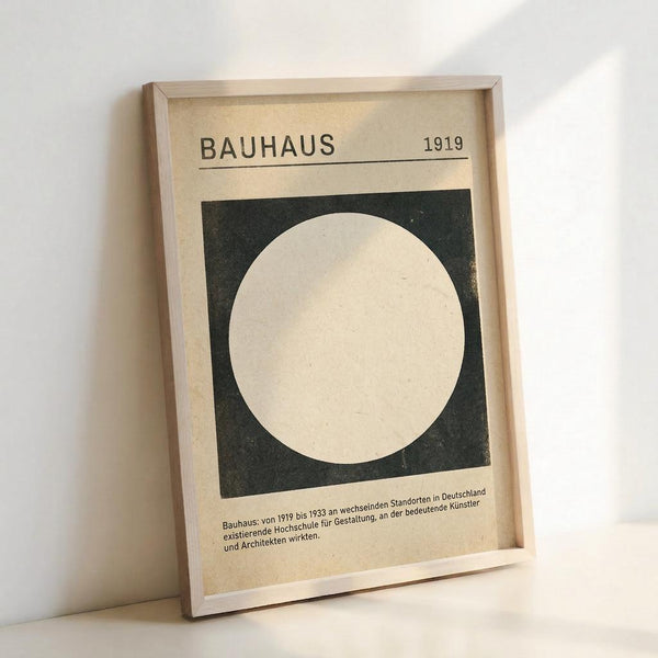 May include: Framed vintage-style Bauhaus poster with cream background featuring bold black square containing white circle, dated 1919, with German text below. Simple geometric design in neutral tones displayed in light wooden frame against white wall.