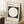 May include: Minimalist Bauhaus-inspired poster with black square containing white circle, displayed in light wooden frame leaning against white paneled wall. German text below describes the design school's history from 1919-1933. Scene shows cozy corner with small wooden side table holding white coffee cup and saucer, textured cream rug, and partial view of woven basket.