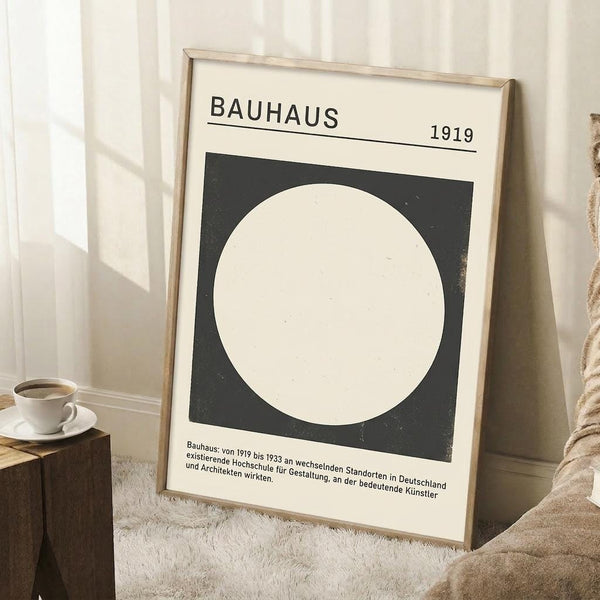 May include: Minimalist Bauhaus-inspired poster with black square containing white circle, displayed in light wooden frame leaning against white paneled wall. German text below describes the design school's history from 1919-1933. Scene shows cozy corner with small wooden side table holding white coffee cup and saucer, textured cream rug, and partial view of woven basket.