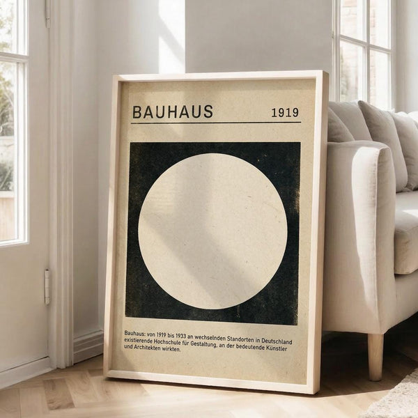 May include: Framed Bauhaus-style minimalist poster featuring a large cream circle on dark green square background, dated 1919, with German text below. Light wood frame leaning against white wall next to beige sofa in bright, modern living room with white wooden floors and large windows.