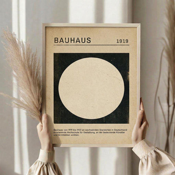 May include: Person in beige clothing holding framed Bauhaus poster against white curtained window. Dried pampas grass visible on left side. Poster features large cream circle on dark green square background with "BAUHAUS 1919" header and German text below. Light wood frame, vintage-inspired design with textured beige background.
