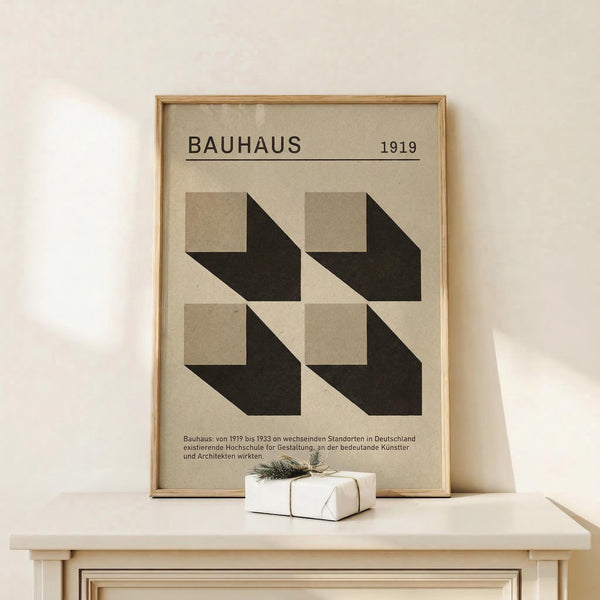 May include: Framed geometric poster with "BAUHAUS 1919" text and four black and beige cube shapes arranged in a grid pattern. Light wooden frame displayed on white wooden console table against cream wall. Small wrapped white gift box with twine and greenery sits beside the frame.