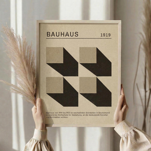 May include: Person holding framed Bauhaus-style geometric poster with four black angular shapes on beige background, dated 1919 with German text below. Light wood frame, neutral beige setting with dried wheat stems visible on left side. Minimalist modernist design print displayed in bright natural lighting.