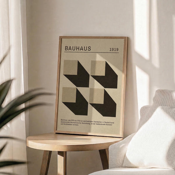 May include: Beige poster with black geometric shapes forming a grid pattern, titled "BAUHAUS 1919" at top with German text below. Framed in thin wood frame, displayed on round wooden side table next to beige upholstered chair. Sunlight streams across white wall creating soft shadows, with green plant leaves visible in corner.