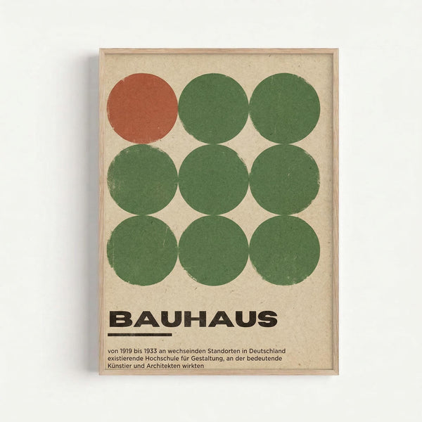 Framed Bauhaus-style poster featuring nine circles arranged in a 3x3 grid on beige textured paper. Eight circles are painted in muted green tones while one circle in the top left is rust orange. Bold black "BAUHAUS" text appears below the circles with German text underneath describing the design school from 1919-1933. The artwork is displayed in a simple light wood frame against a white wall.
