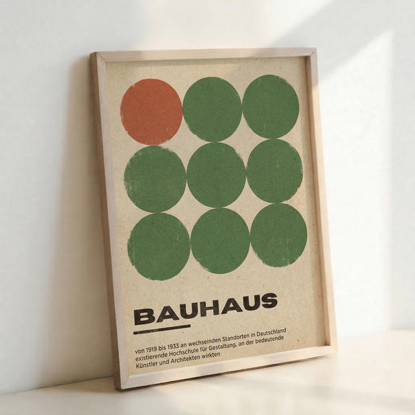 May include: Framed Bauhaus poster print with nine painted circles arranged in a 3x3 grid - one rust-colored circle in top left, eight sage green circles filling remaining positions. Black "BAUHAUS" text below with German descriptive text. Light wood frame on beige textured paper background, leaning against white wall with subtle shadows.