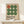 May include: Framed Bauhaus-style poster with nine circles arranged in a 3x3 grid - one rust-colored circle in top left, eight green circles in varying shades. Text reads "BAUHAUS" with German description below on cream background. Natural wood frame sits on white wooden shelf against white wall, with small wrapped gift box nearby.