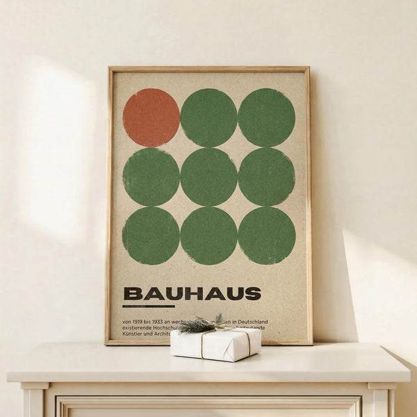 May include: Framed Bauhaus-style poster with nine circles arranged in a 3x3 grid - one rust-colored circle in top left, eight green circles in varying shades. Text reads "BAUHAUS" with German description below on cream background. Natural wood frame sits on white wooden shelf against white wall, with small wrapped gift box nearby.